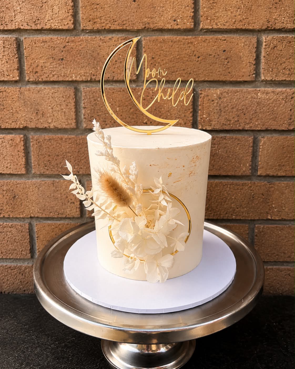 Ivory Moon Child birthday cake with gold Moon Child acrylic topper, dried pampas grass, white hydrangeas, eucalyptus and gold hoop detail
